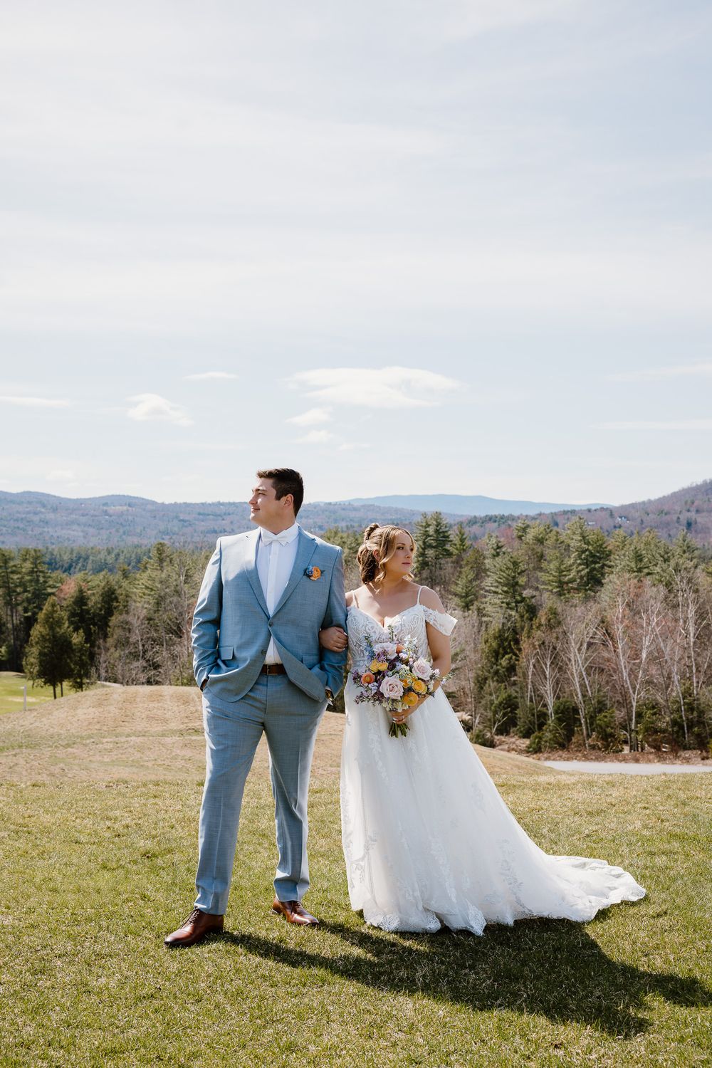 Bride and groom pose on a scenic hilltop with mountain views, she in a lace gown holding a bouquet, he in a blue suit.