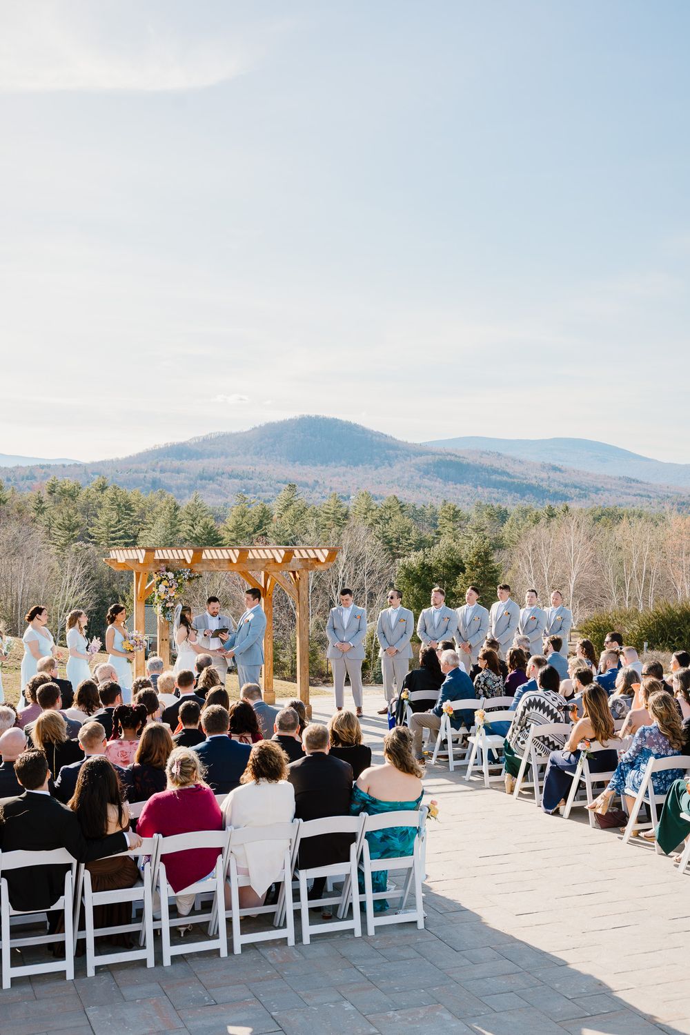 Outdoor wedding ceremony on a stone terrace with mountain views, wooden pergola altar, and guests seated in white chairs.