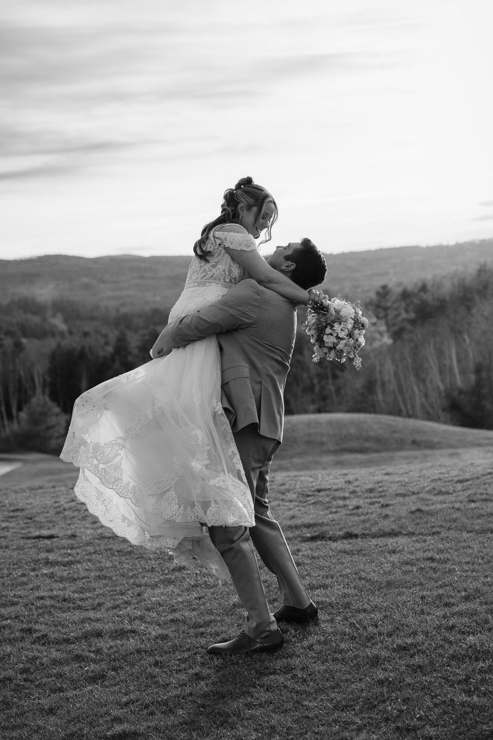 Black and white photo of groom lifting bride in wedding dress with bouquet in scenic outdoor setting.