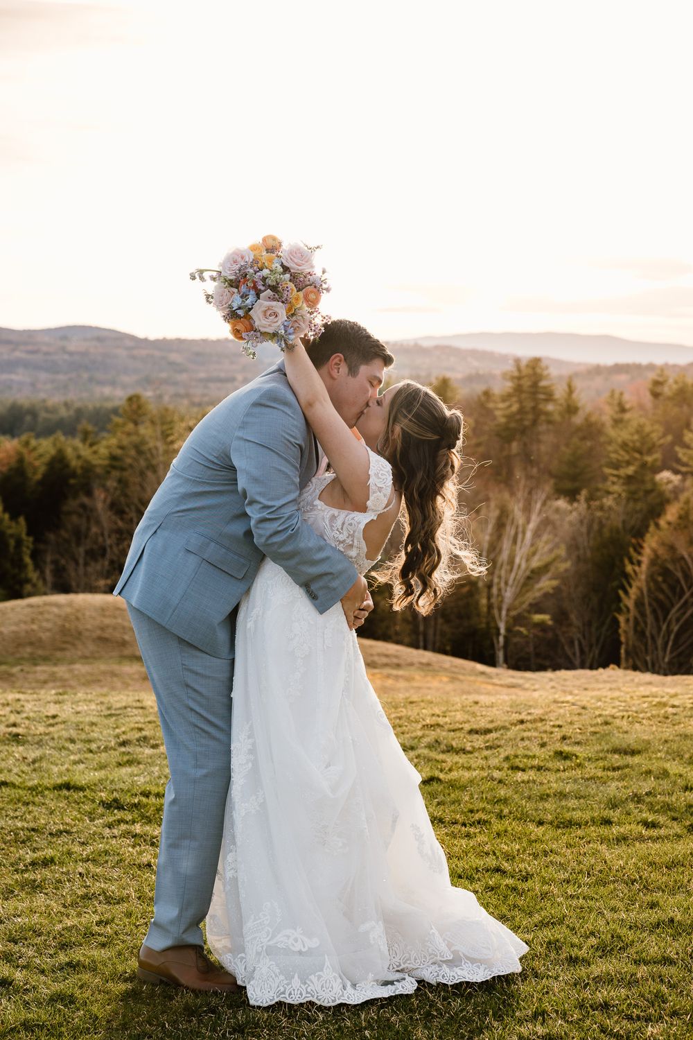 Bride and groom share a romantic dip kiss at sunset on a green hillside, with mountain views and a colorful bouquet raised high.