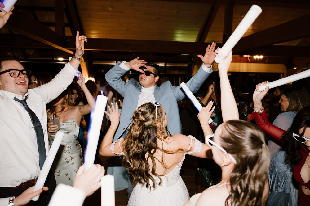 Bride and groom dancing with wedding guests holding glow sticks at a lively indoor wedding reception celebration.