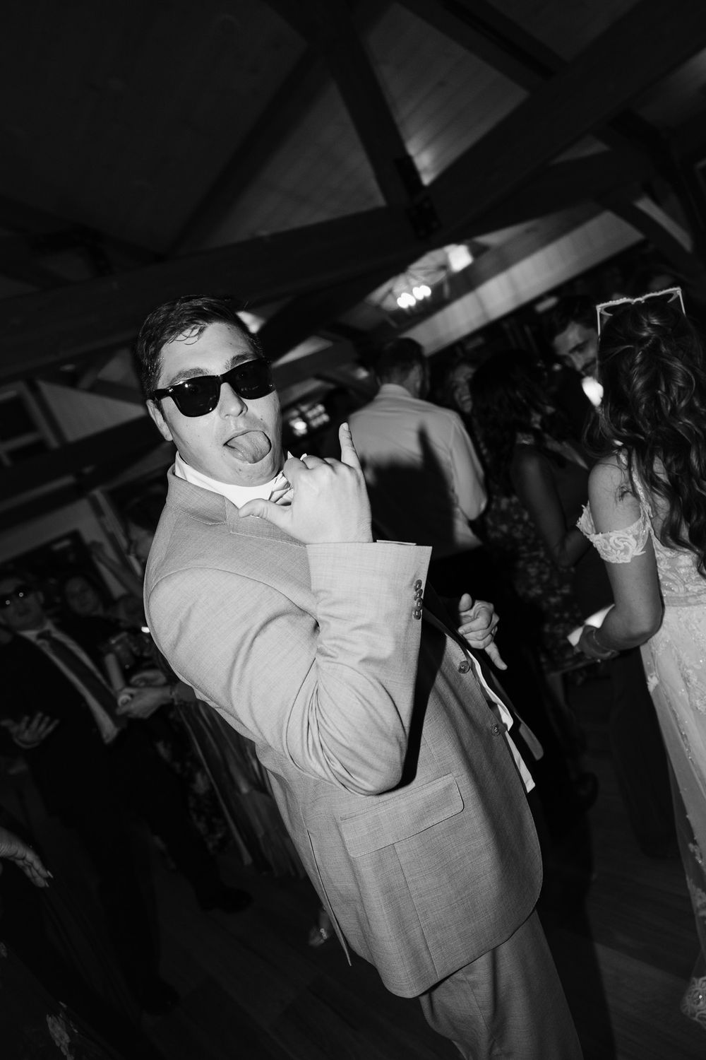 Groom in a suit and sunglasses strikes a fun pose on the dance floor at a lively wedding reception.