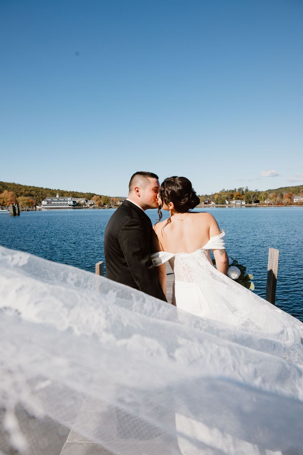 Bride and groom share a kiss on a lakeside dock, bride in off-shoulder gown with flowing veil, clear blue sky backdrop.