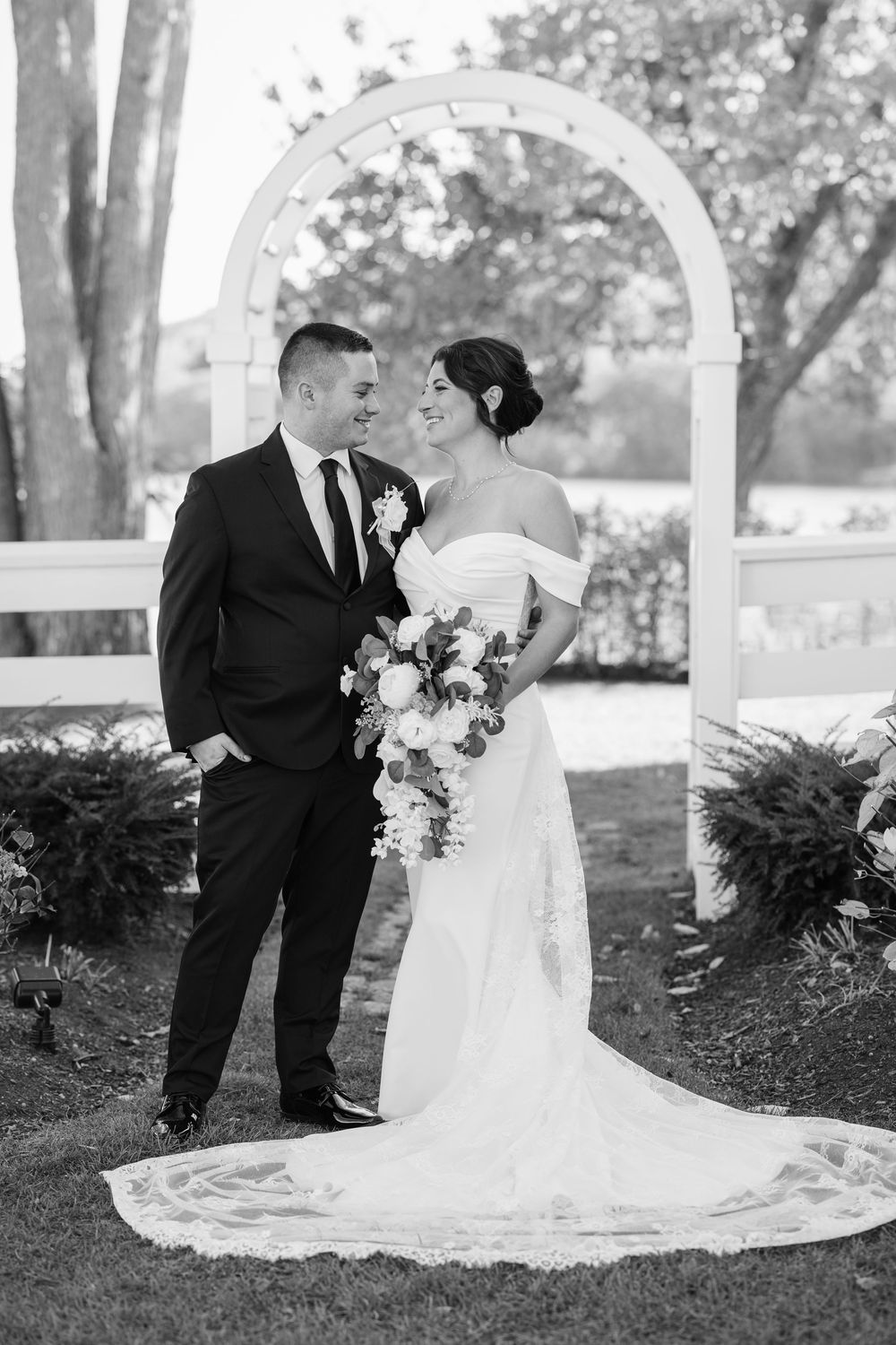 Black and white wedding photo of bride and groom standing under white arch, bride holding bouquet in elegant gown.