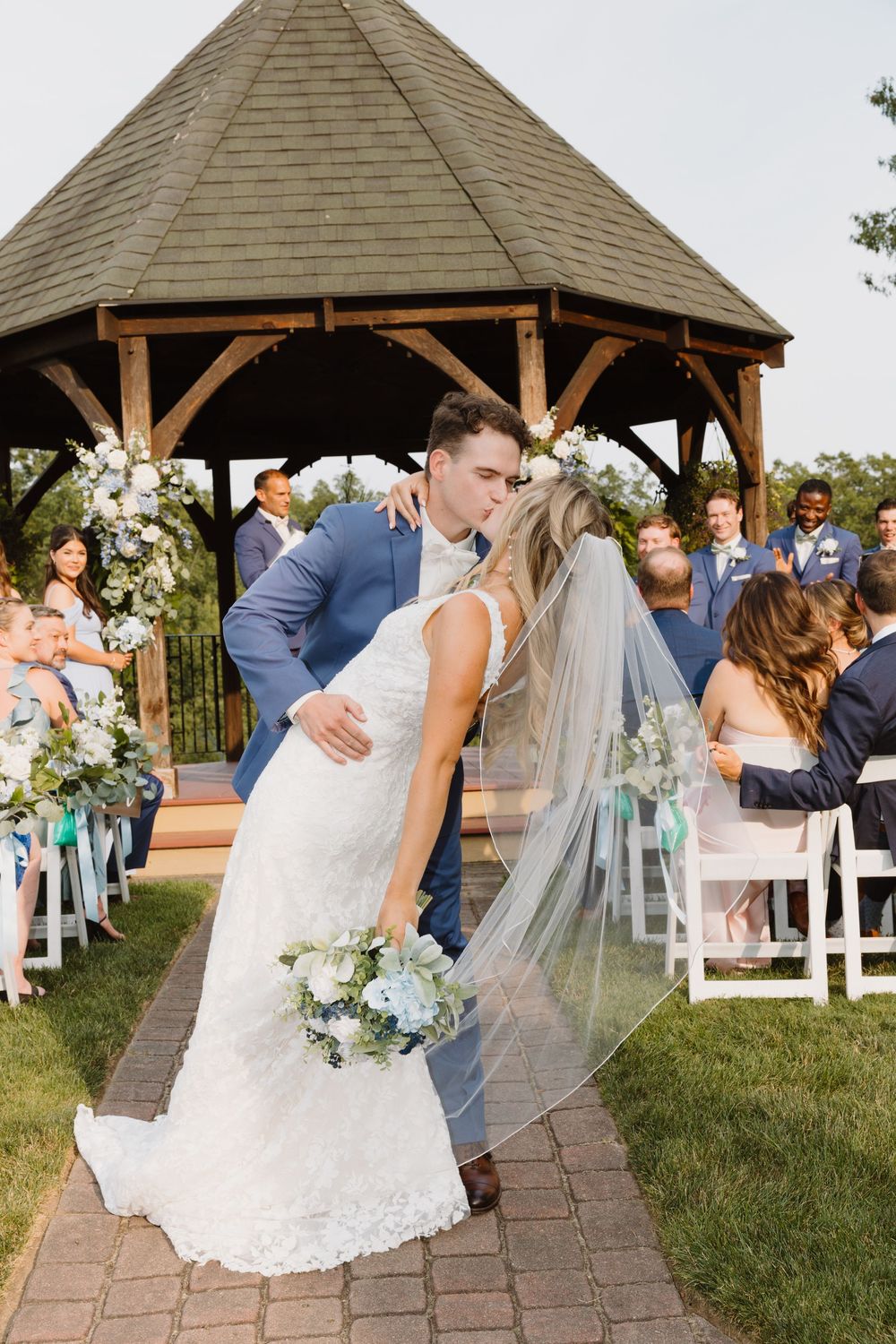 Newlyweds share their first kiss under a rustic outdoor gazebo surrounded by guests and floral decorations.