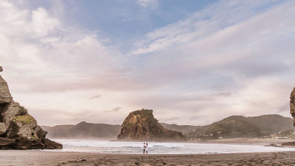 Dramatic Piha coastal engagement shoot couple landscape with rock formations and misty beach at sunset.