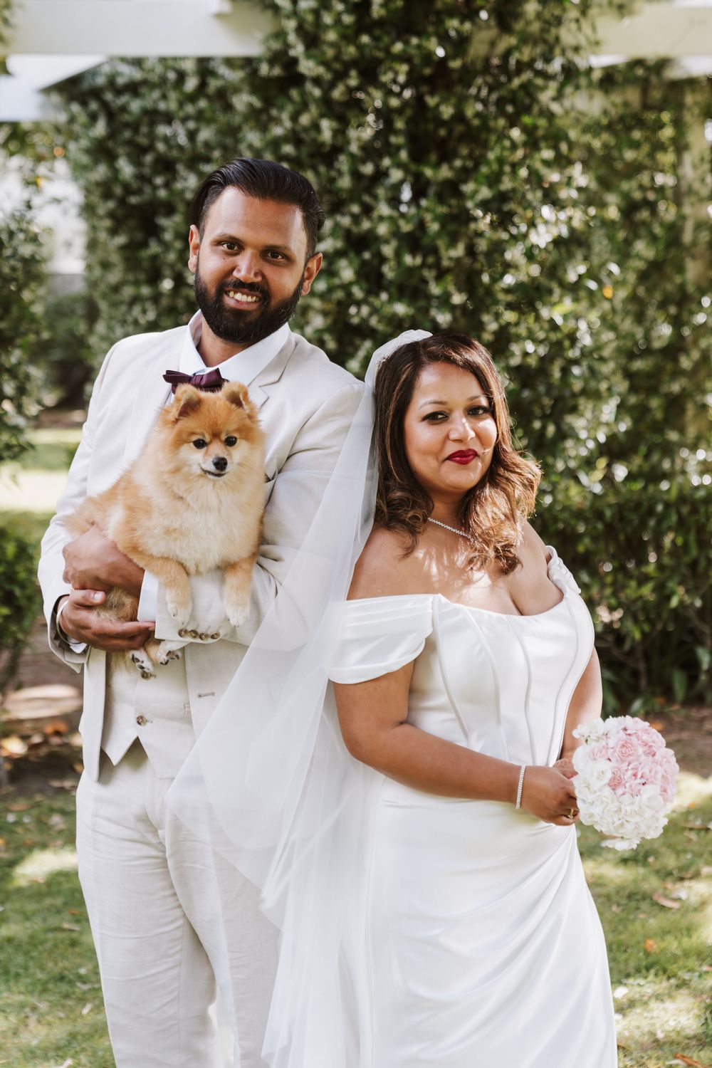 Wedding couple in white attire poses with their adorable Pomeranian dog in a garden setting.