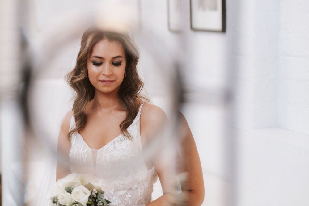 Bride in white lace dress holding bouquet during wedding preparations.