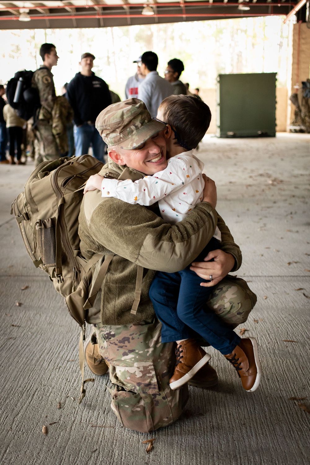 A smiling soldier in full gear kneels to hug a young child during a military homecoming.
