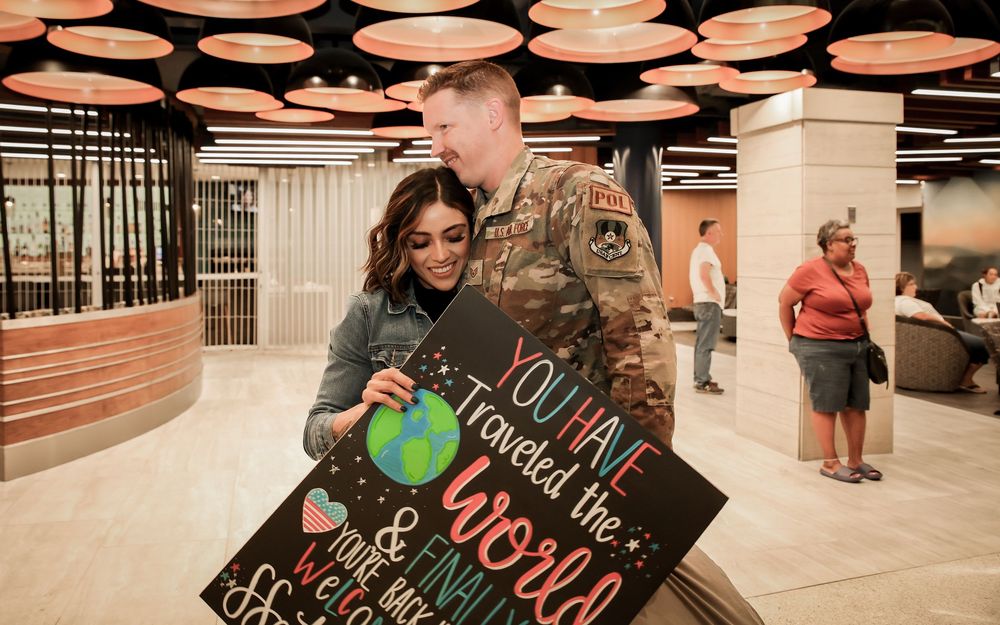 A soldier in uniform embraces a young woman holding a welcome home sign at an airport.