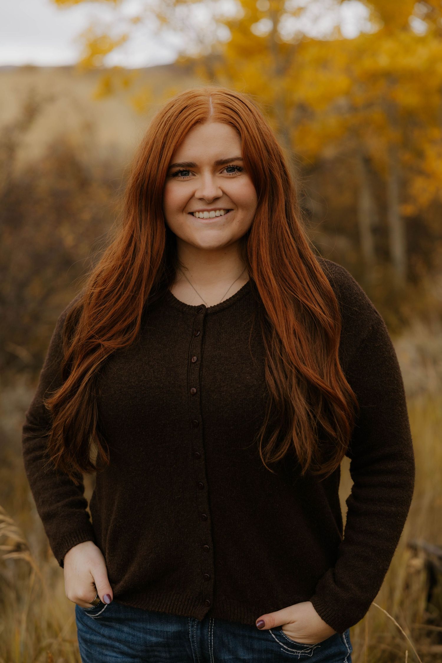 Woman in denim and brown vest poses in autumn field with mountains in background.