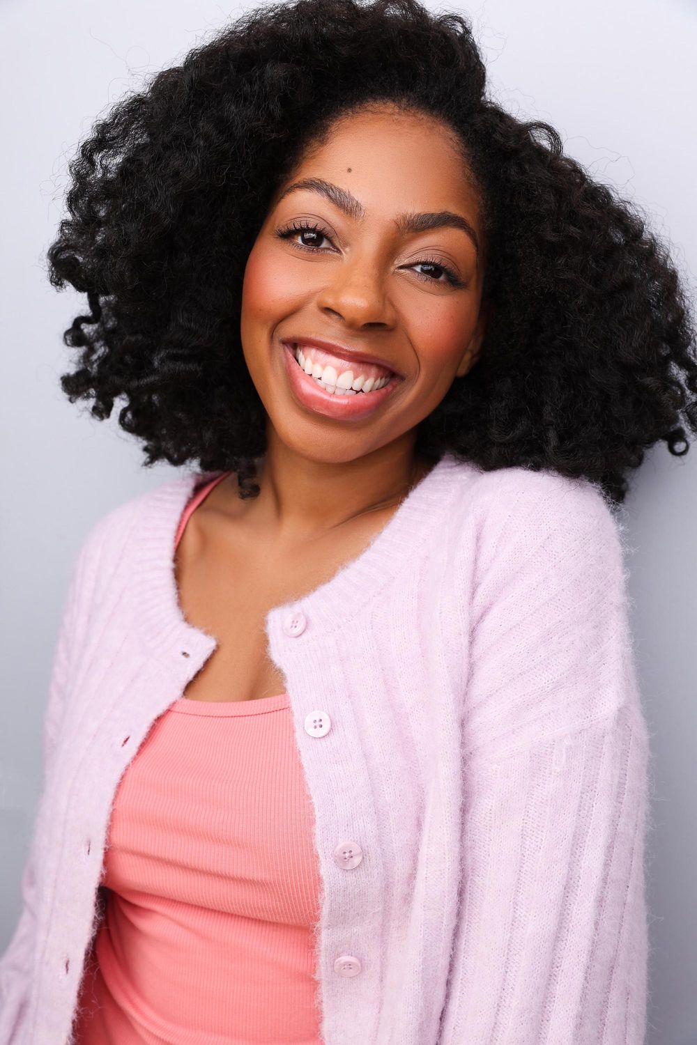 A smiling Black woman with curly hair wears a pink cardigan over a coral top against a gray background.