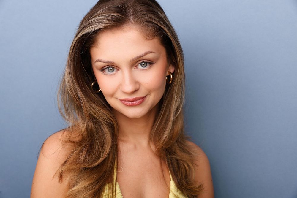A young woman with long brown hair and blue eyes smiles softly wearing a gold top against a blue background.