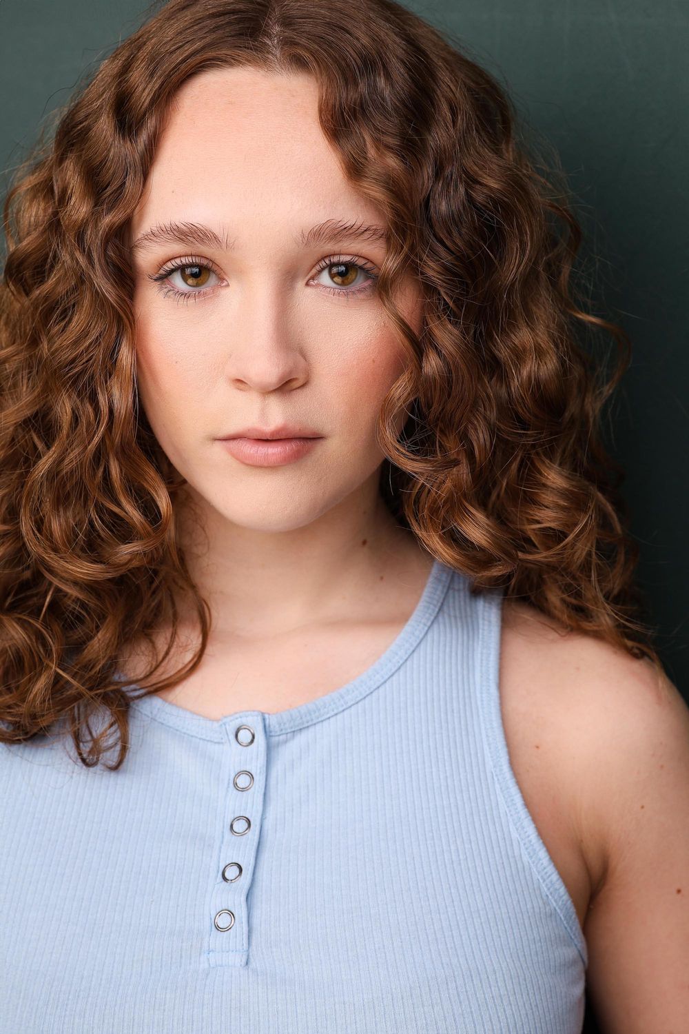 Young woman with curly auburn hair wearing a light blue ribbed tank top poses for a headshot photo.