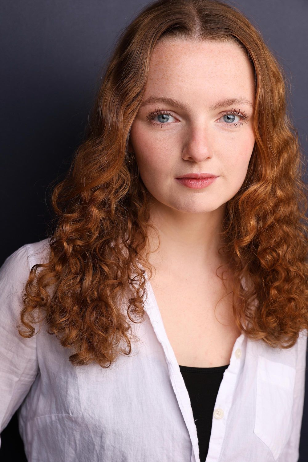 Young woman with curly red hair and blue eyes wearing a white shirt against a gray background.