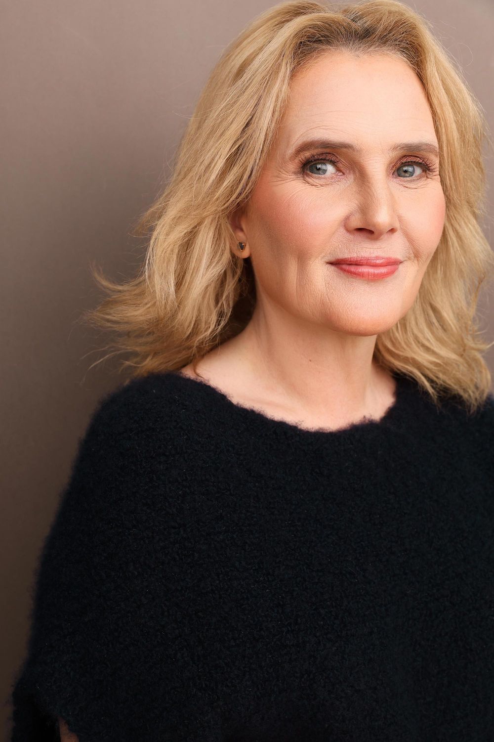 Elegant blonde woman with blue eyes wearing a black sweater against a taupe background.