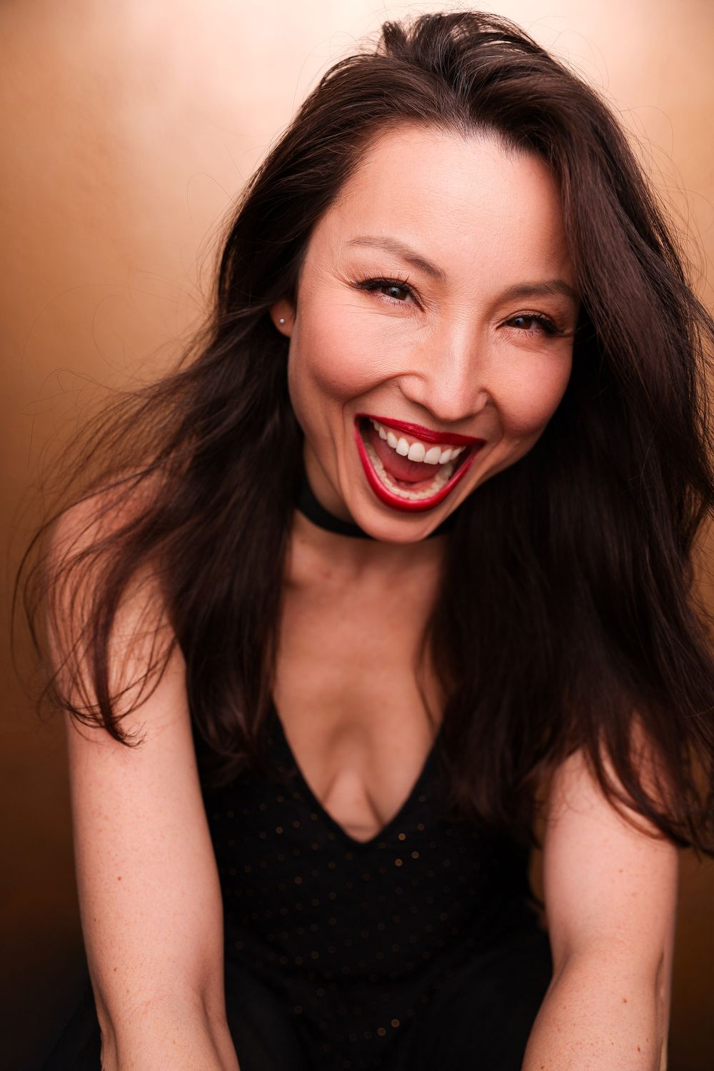 Joyful Asian woman with long dark hair and red lips laughing, wearing a black top on a gold background.