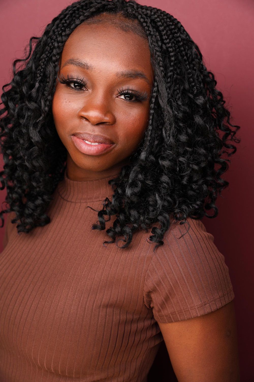 Young Black woman with box braids and curly ends wearing a brown ribbed top, posing against a dark red background.