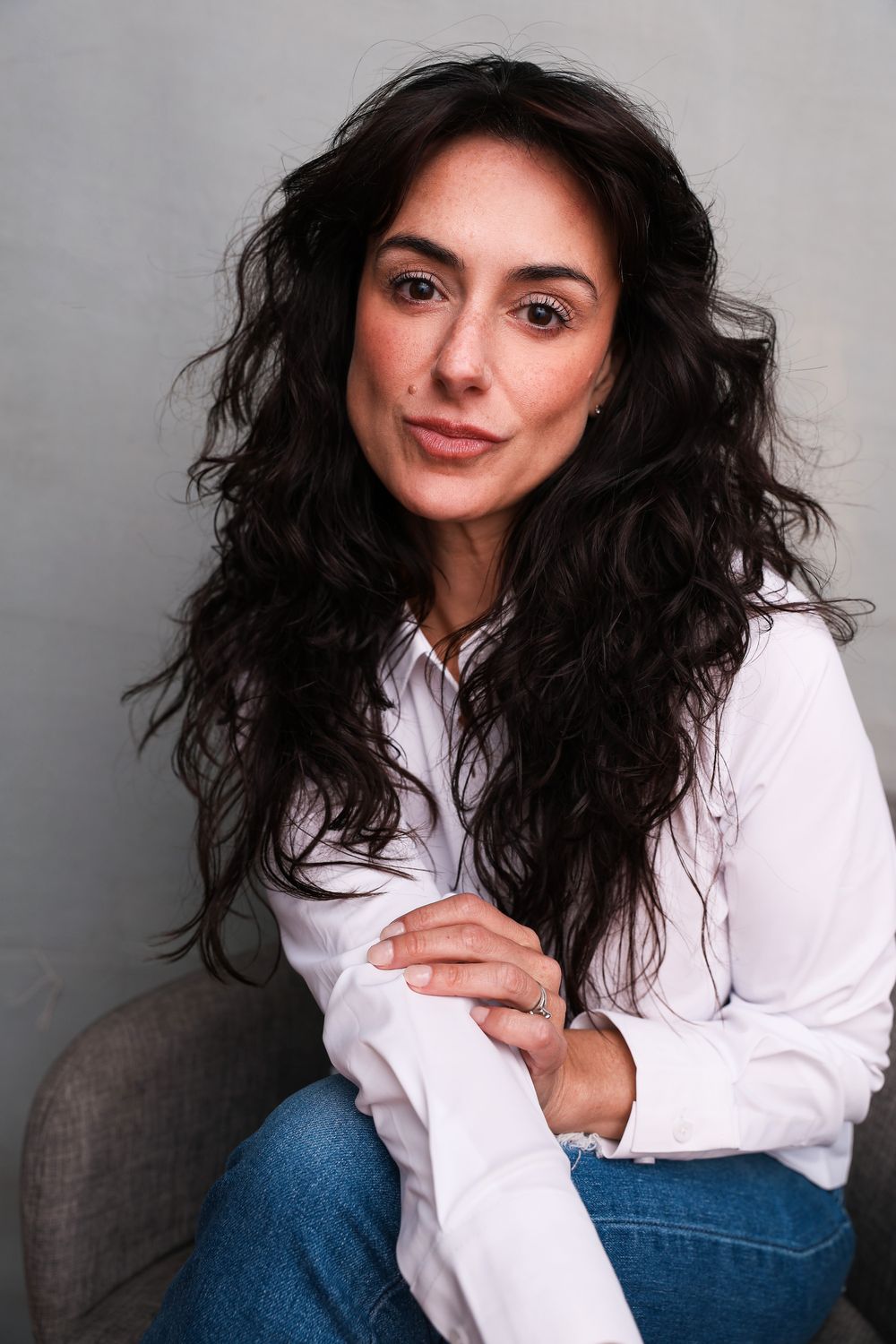 Woman with long dark curly hair wearing white shirt and jeans, sitting relaxed against gray background.