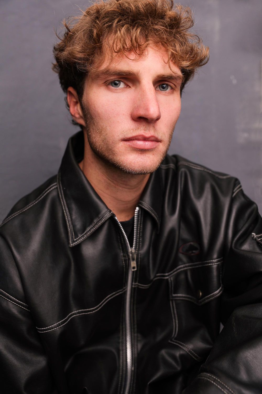 Young man with curly hair wearing black leather jacket posing against gray background.