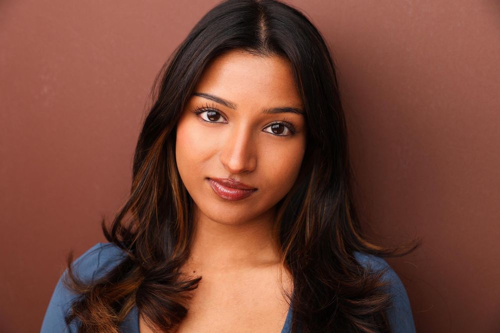 Young South Asian woman with long dark wavy hair wearing a blue top, posing against a brown background.