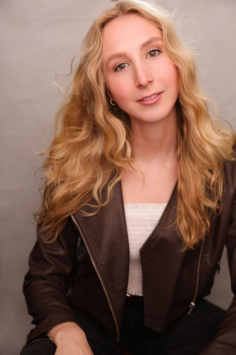 Blonde woman in brown leather jacket with wavy hair smiling softly against gray background.