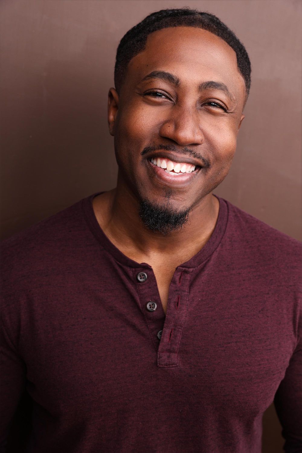 Smiling Black man with beard wearing a maroon henley shirt poses for a headshot against a brown background.