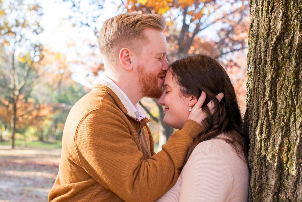Couple sharing a romantic moment leaning against a tree in a colorful fall park.