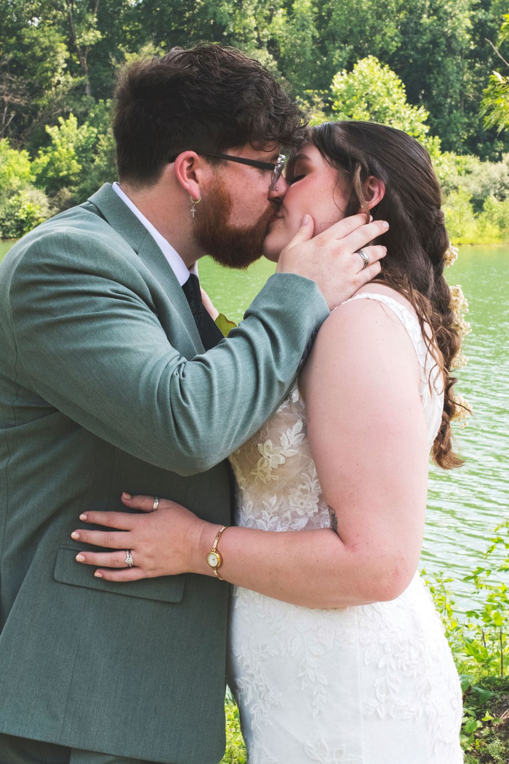 Newlywed couple shares their first kiss outdoors near a scenic green pond on their wedding day.