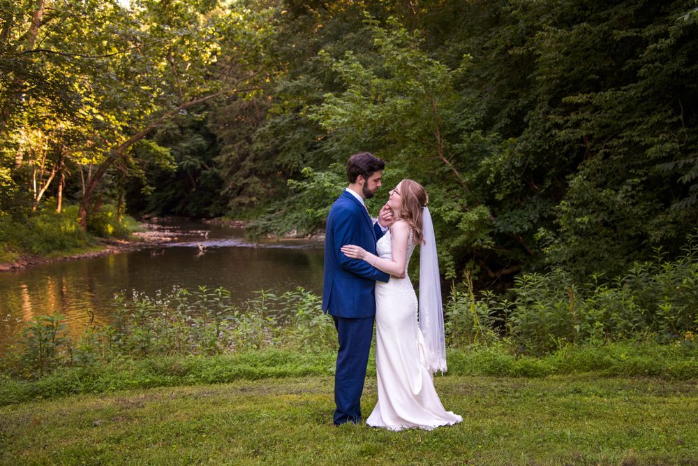 Romantic bride and groom embrace near a peaceful pond surrounded by beautiful green trees.