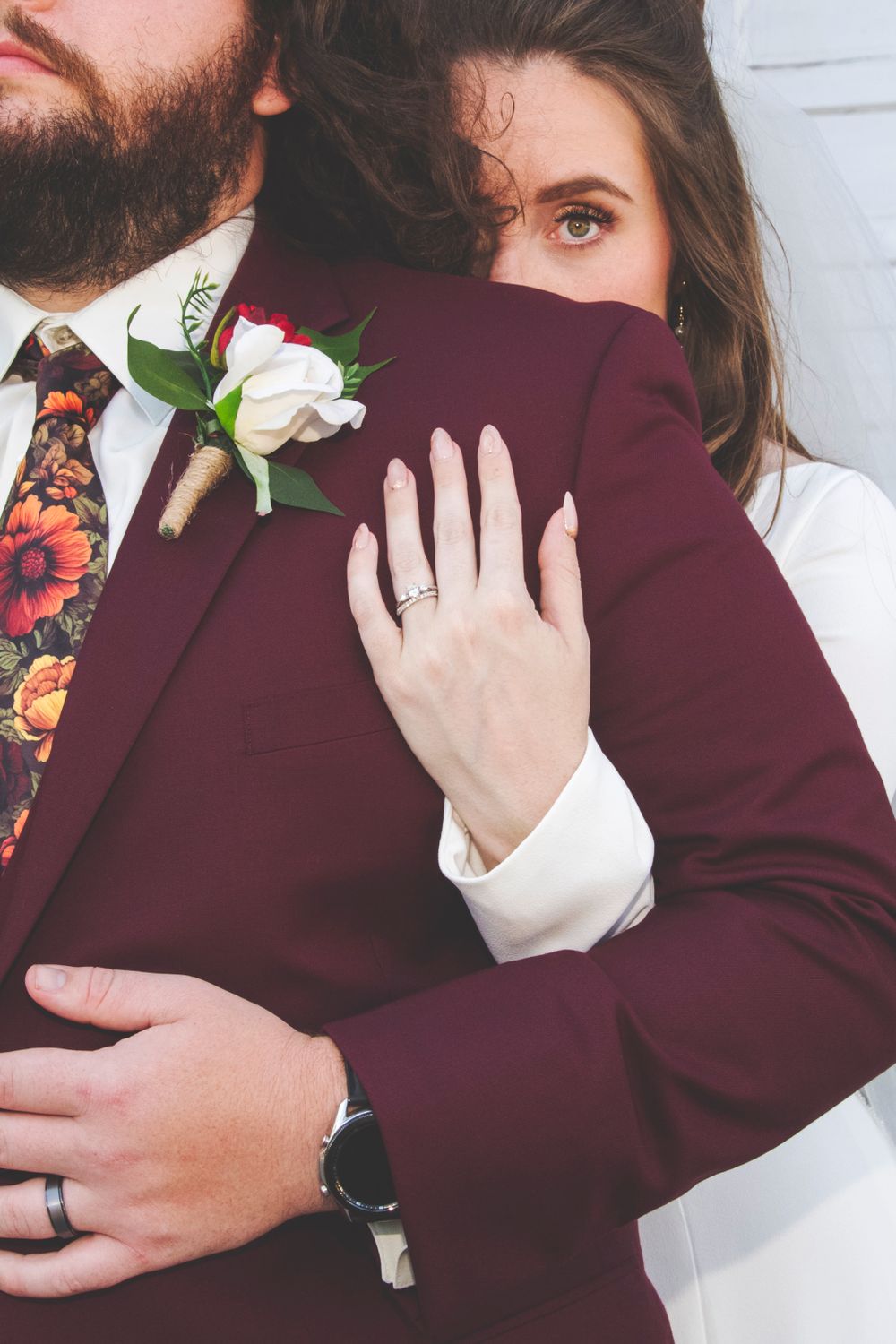 Bride resting her head on groom's burgundy suit, showcasing wedding rings and floral boutonniere.