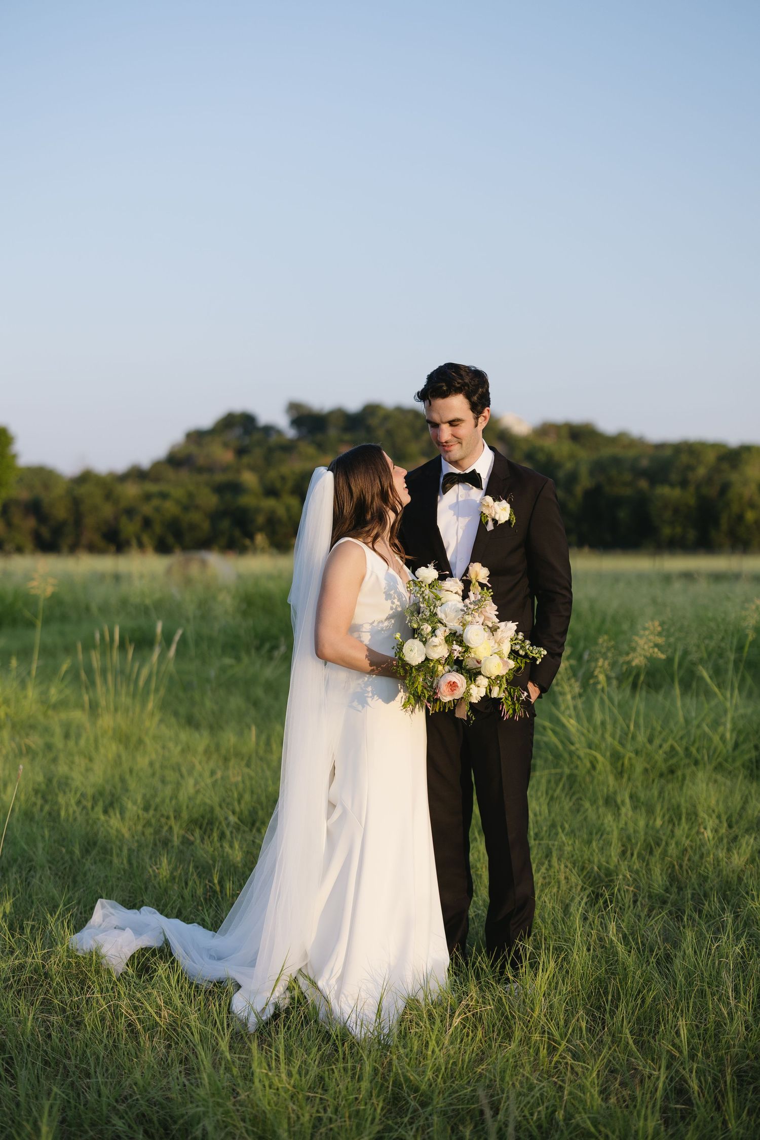 Bride and groom in formal attire standing in a lush green field at golden hour with bouquet.
