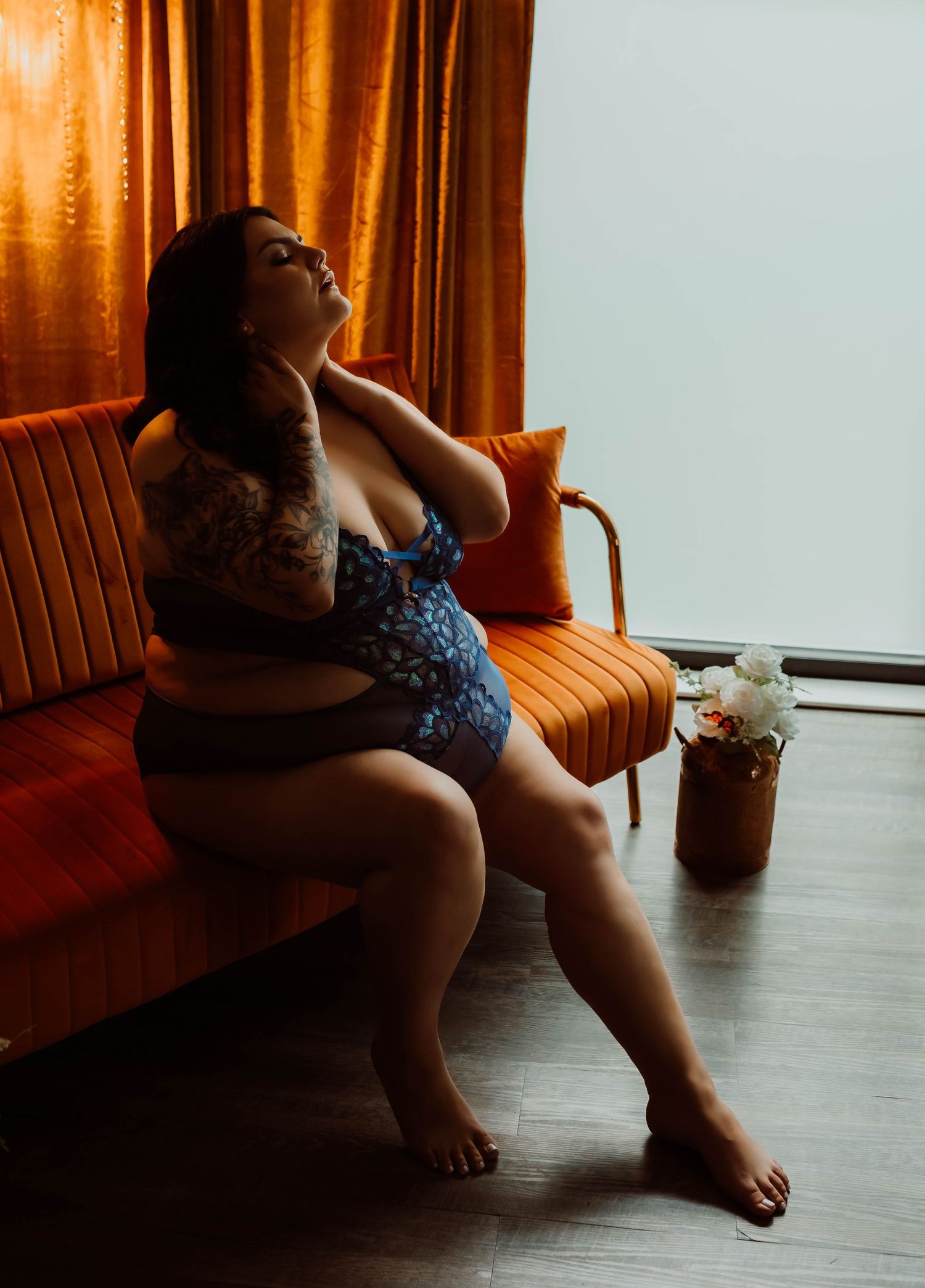 A woman in lingerie sits on an orange vintage couch near a window with warm sunlight streaming through curtains.