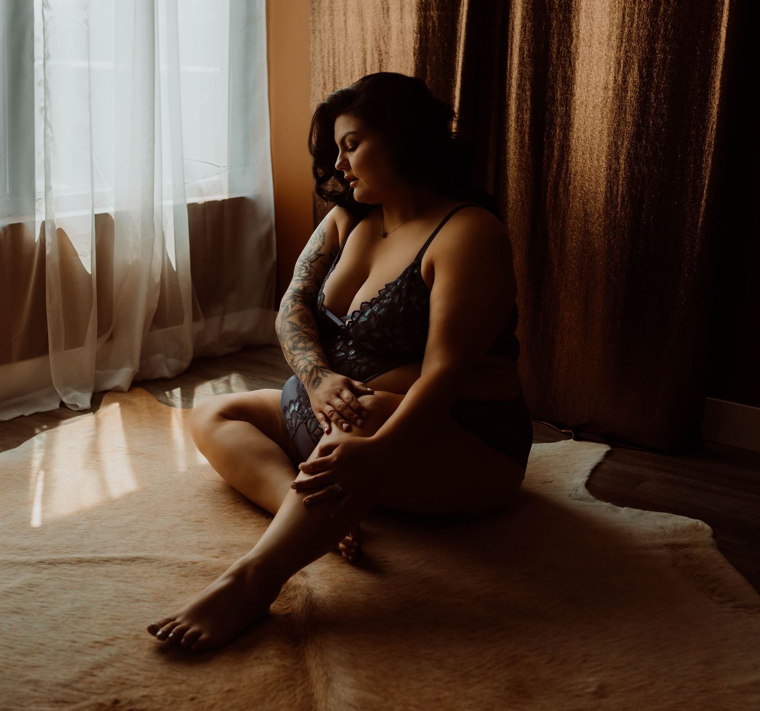 A silhouette of a women sits near curtained windows in a moody, atmospheric room with soft natural lighting for her boudoir photoshoot.
