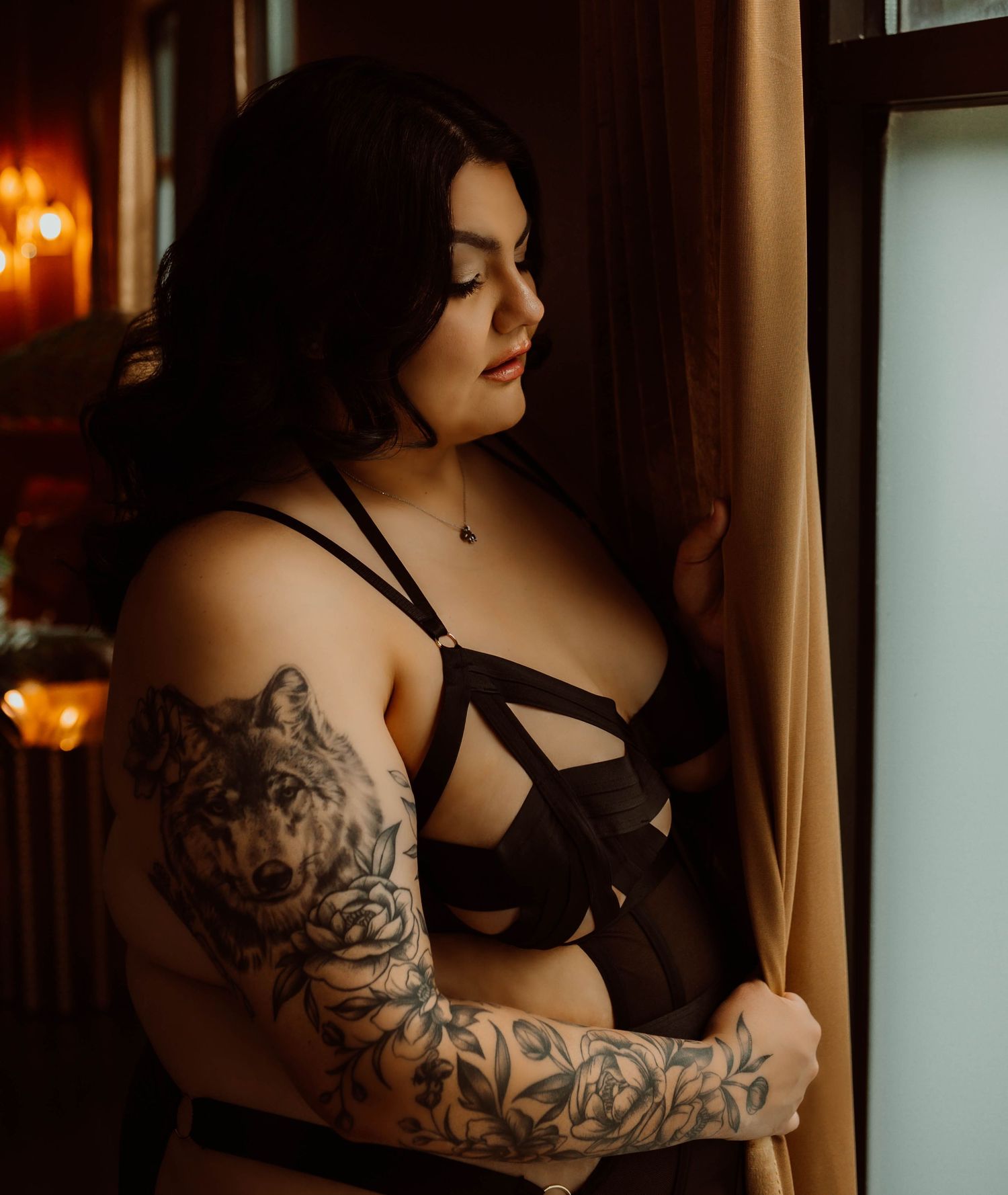 A women in black lingerie poses artistically by a window with dramatic lighting and visible arm tattoos for boudoir photos.