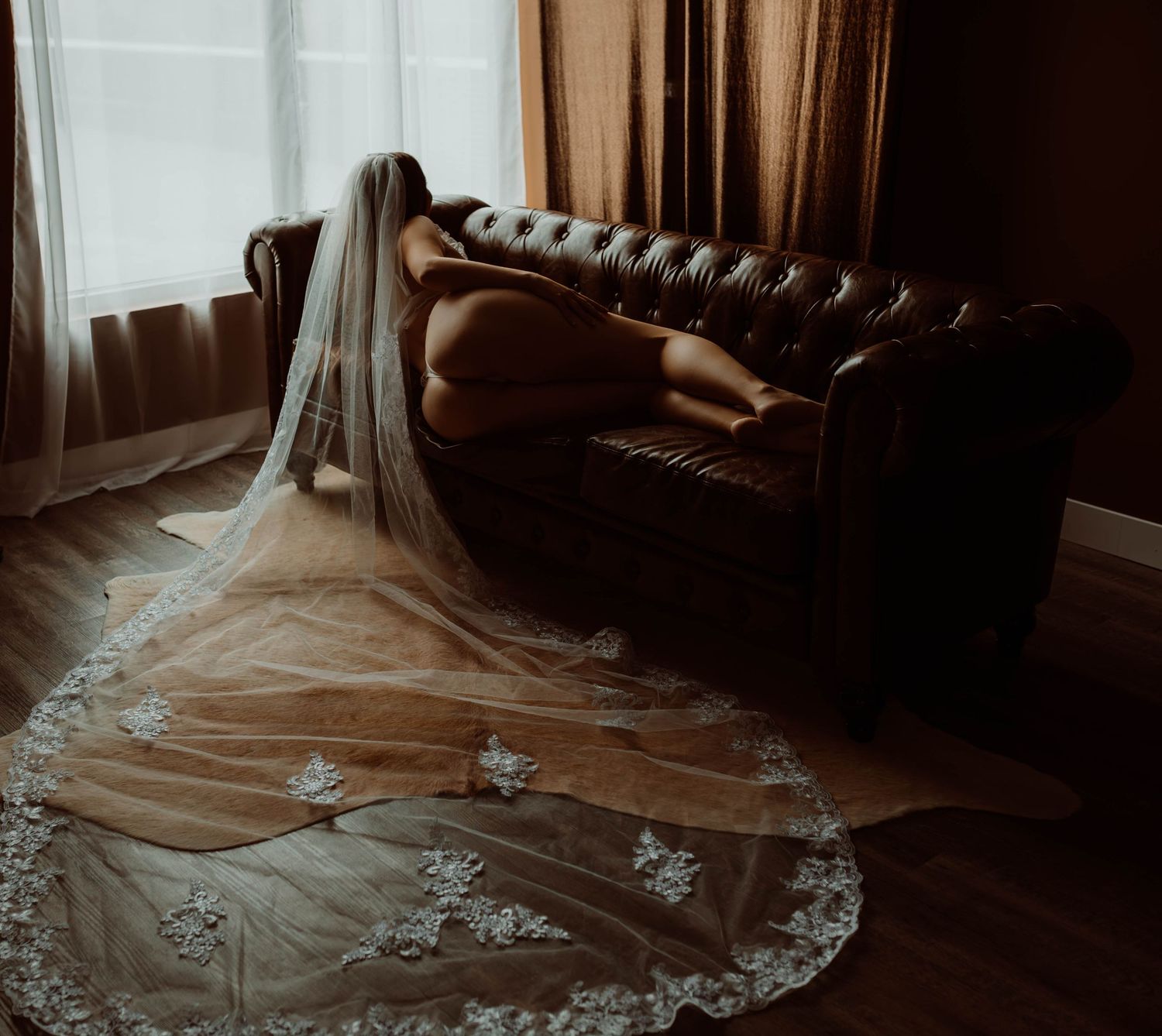 Bridal boudoir photography delicate lace wedding veil drapes elegantly over a brown leather chesterfield sofa in moody lighting.