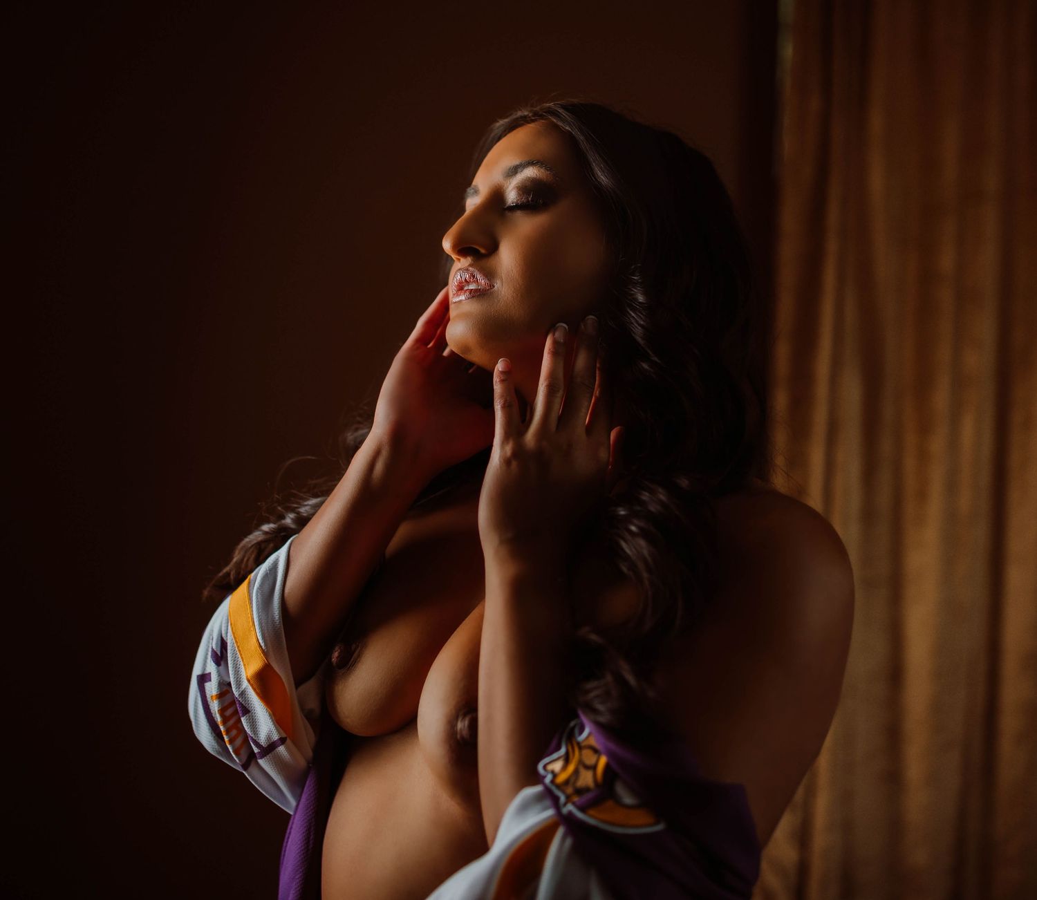 A woman poses gracefully in soft warm light, with a football jersey, with hands framing her face during a artistic boudoir photoshoot.