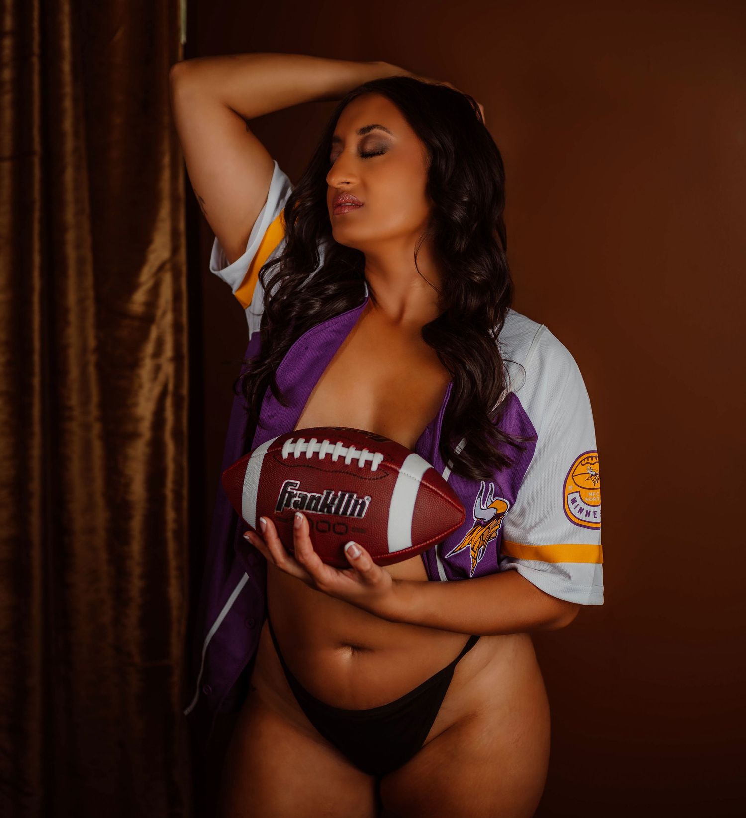 Boudoir photoshoot of a woman in a Minnesota Vikings jersey holding a football in a moody boudoir studio.