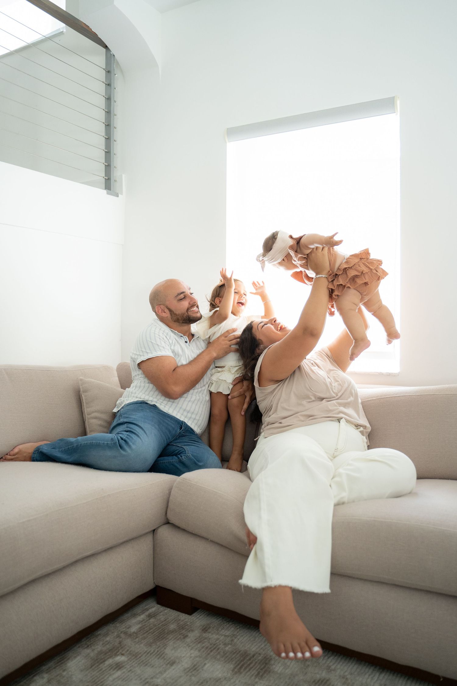 Happy family playing with baby on a cozy beige sofa in a bright modern living room.