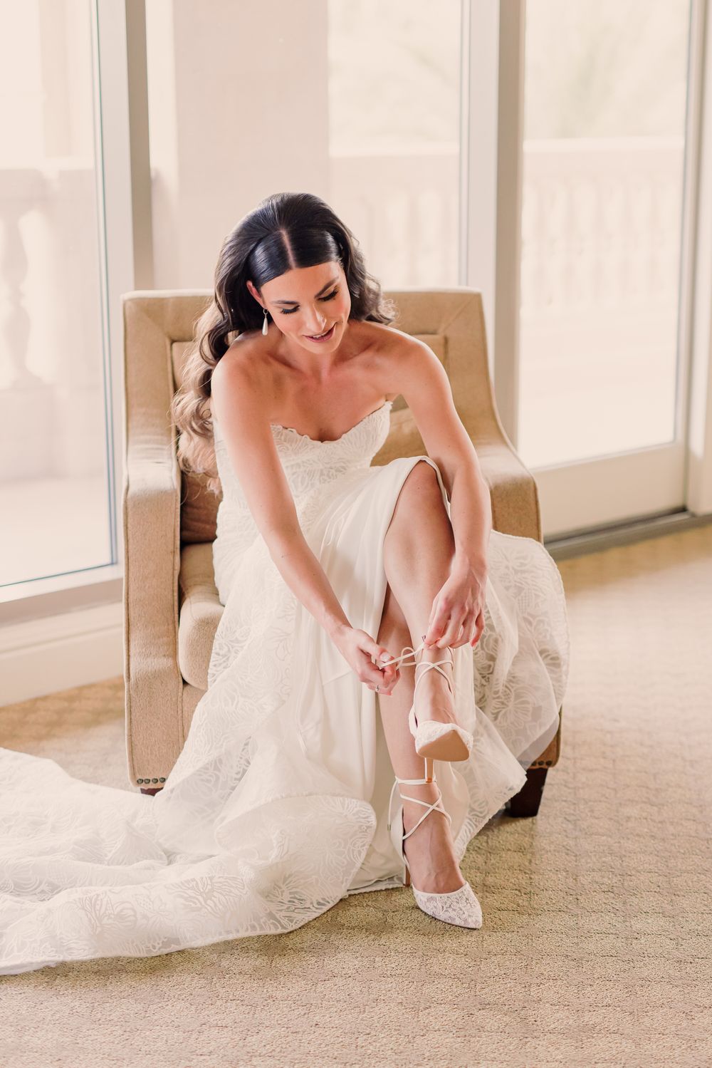 A bride in a white wedding gown sits on the floor adjusting her elegant bridal shoes in a sunlit room.