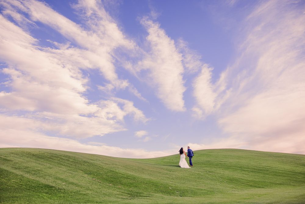 A bride and groom walk along rolling green hills under dramatic clouds at sunset.