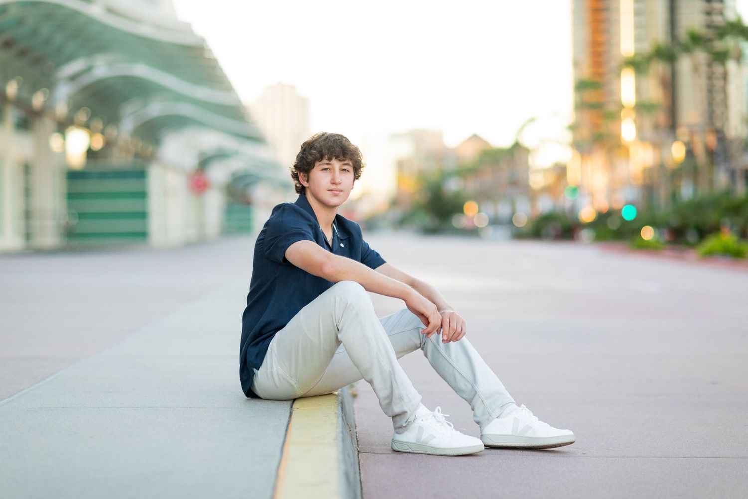 A high school senior in casual attire sits casually on a curb during sunset with modern architecture in the background.