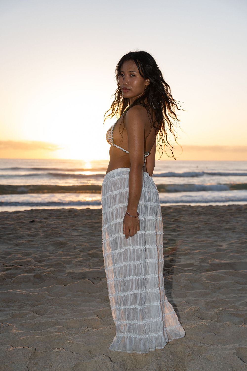 Woman in bikini top and white maxi skirt posing on beach at golden sunset with ocean waves behind her.