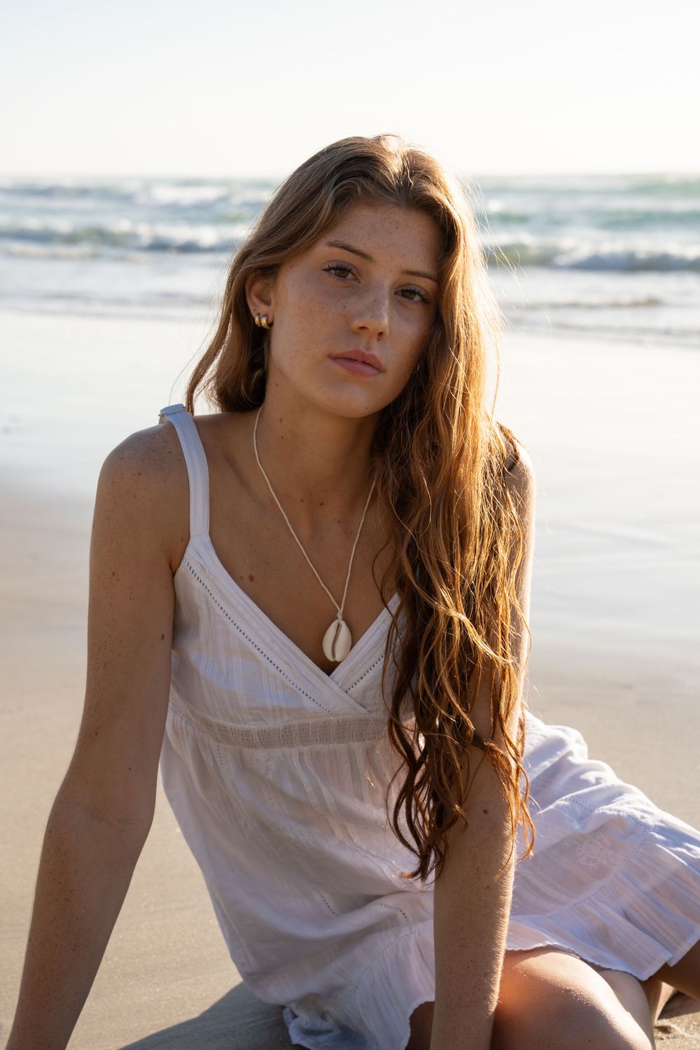 Young woman in white sundress sitting on sandy beach with ocean waves, wearing shell necklace, golden hour light.