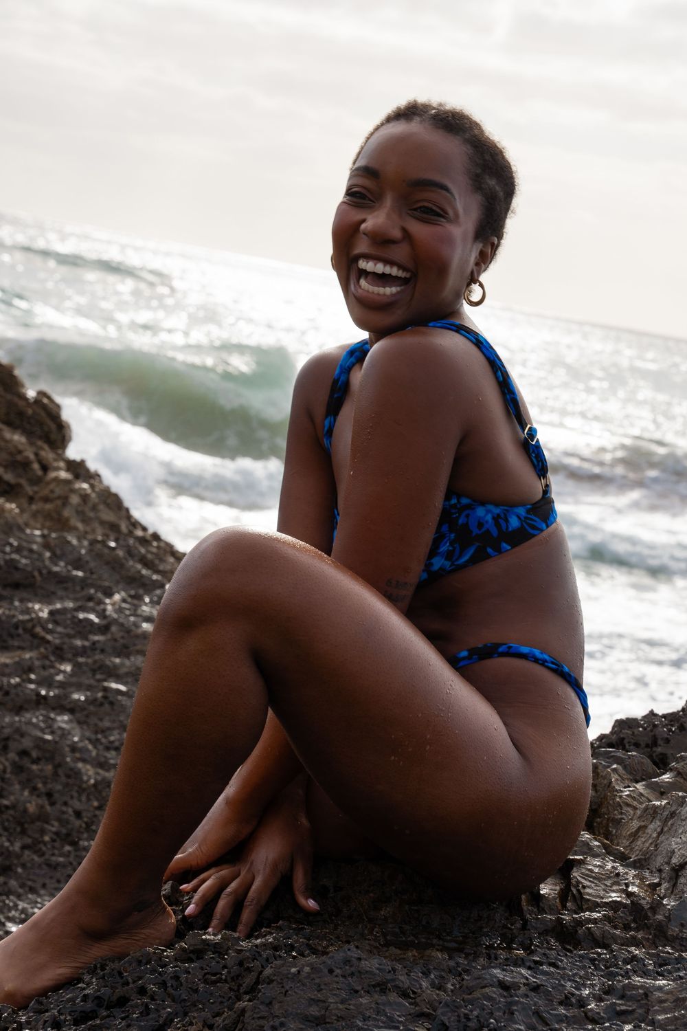 A smiling woman in a blue patterned bikini sits on dark rocks by the ocean with waves in the background.