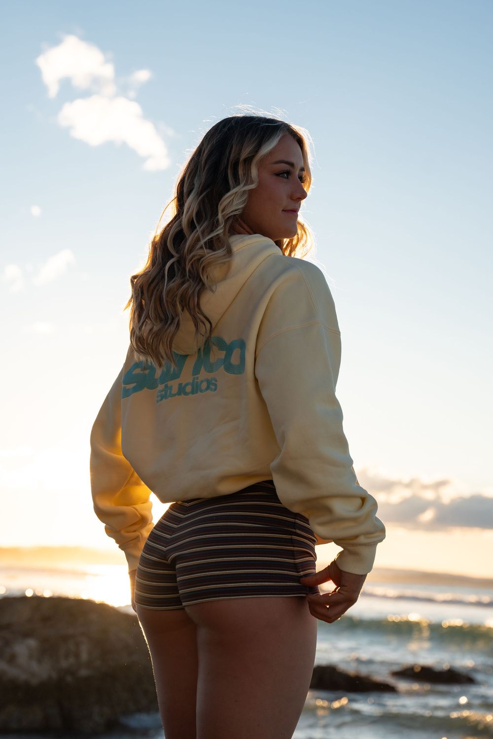 Blonde woman wearing yellow surf studio hoodie and striped shorts standing on rocky beach at golden hour.