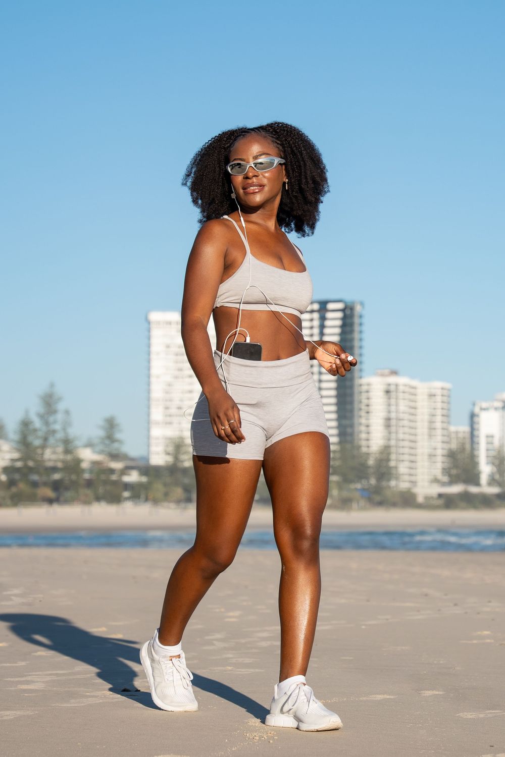 A fit Black woman in beige athletic wear and sunglasses walks on a sunny beach with city buildings in the background.