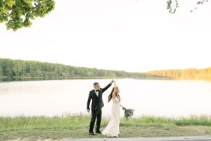Newlyweds holding hands and walking by a serene lake at sunset.