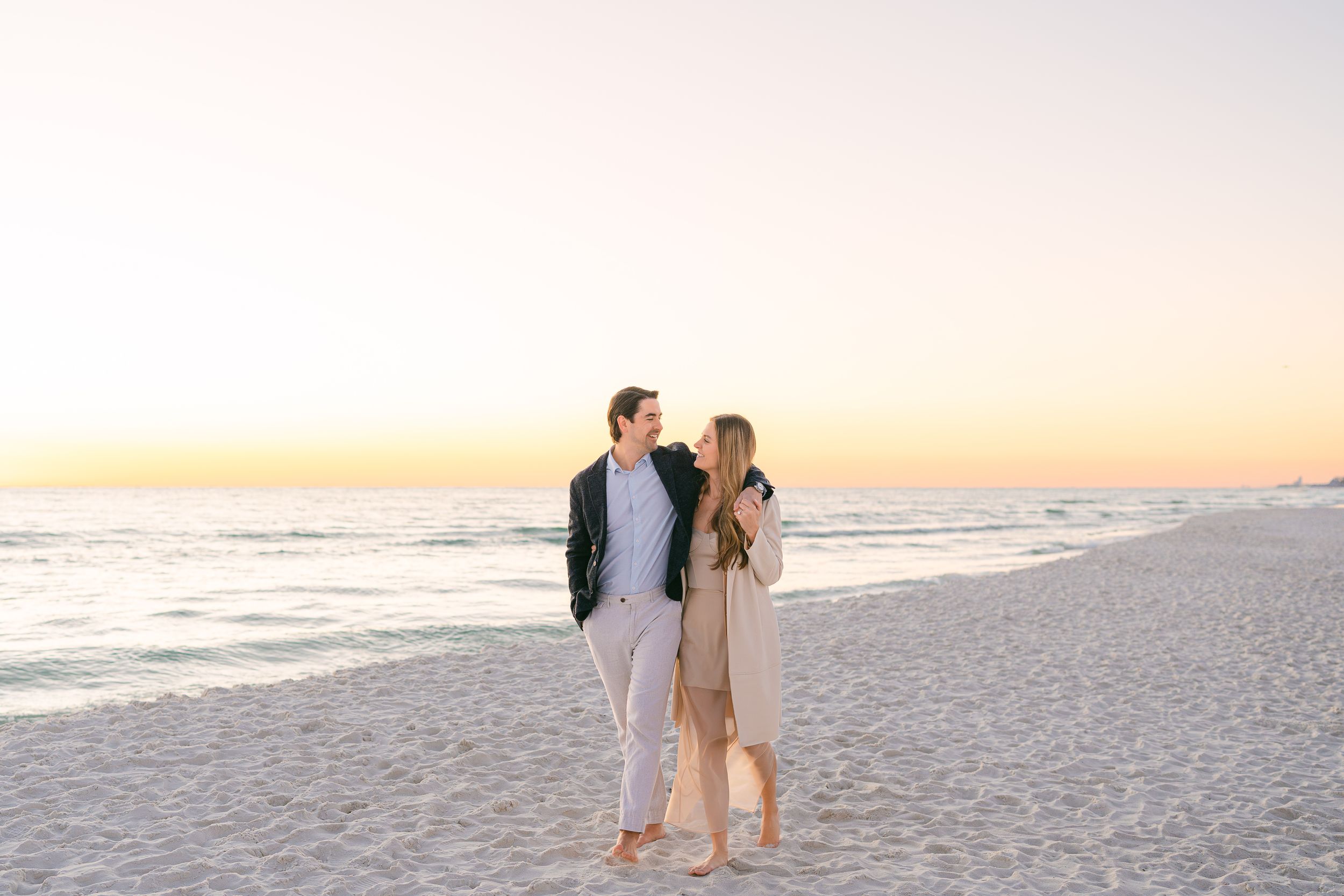 A couple shares a romantic moment while walking on a sandy beach at sunset with ocean waves in the background.