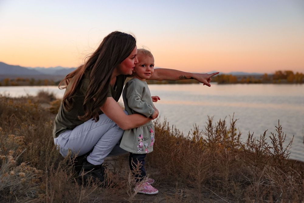 A touching moment between parent and child pointing at something near a lake at sunset.